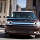 A photo of the front of a Ford Flex driving through a city.